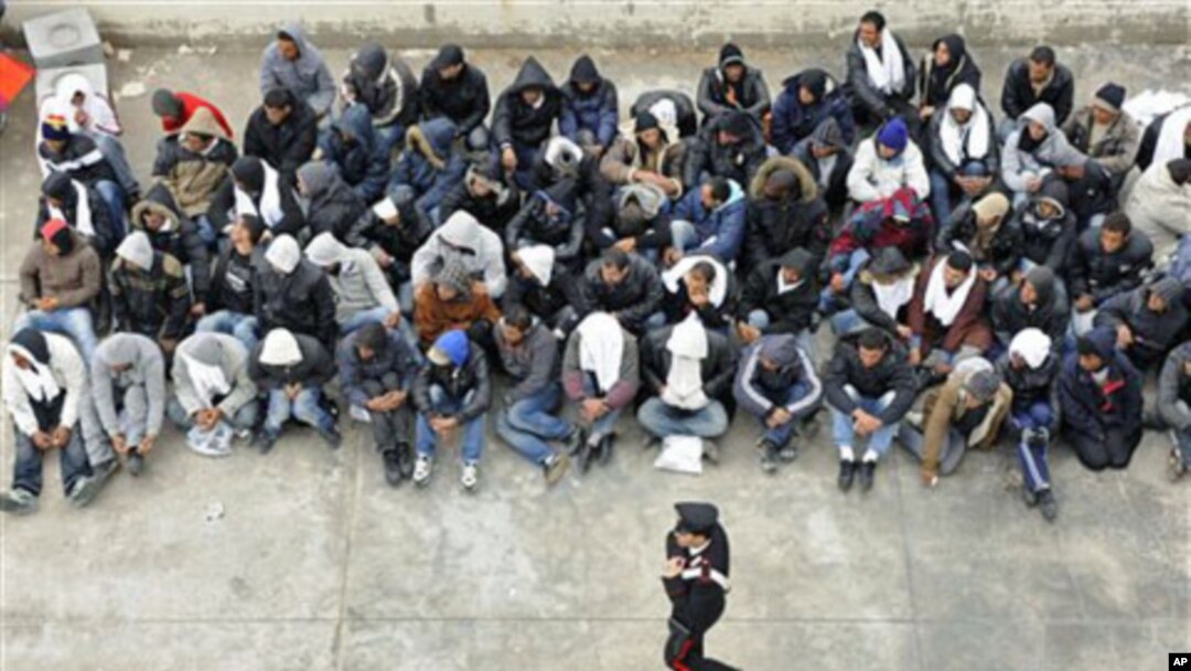 A Carabinieri police officer walks past Tunisian migrants as they wait to board a ship towards Porto Empedocle, in Sicily, where they will be taken for document checks, in Lampedusa, Italy, February 14, 2011