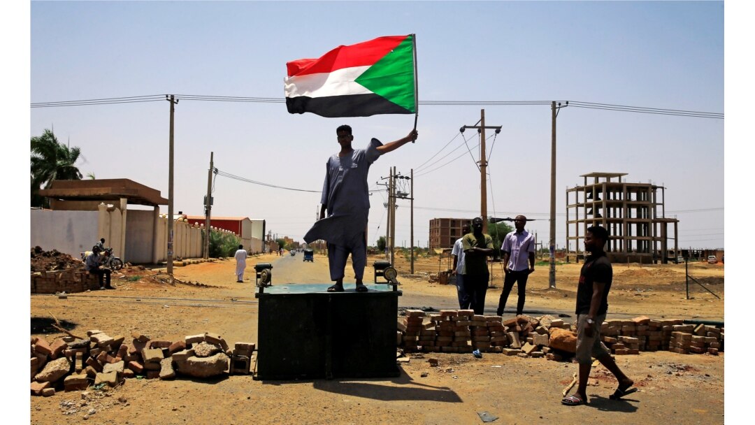 FILE - A Sudanese protester holds a national flag as he stands on a barricade along a street, demanding that the country's Transitional Military Council hand over power to civilians, in Khartoum, Sudan, June 5, 2019.