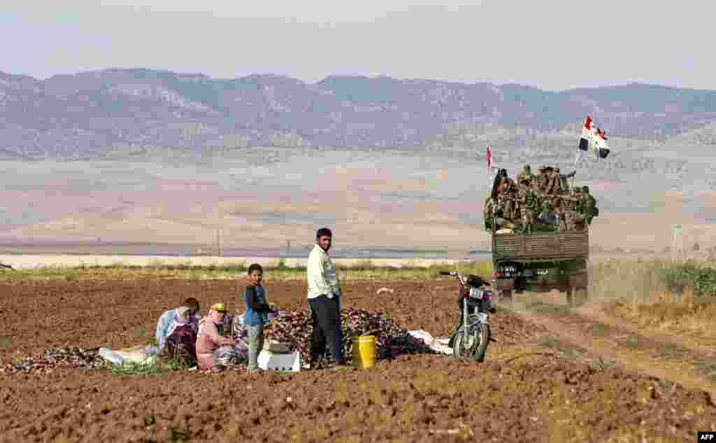 Syrian government soldiers ride in the back of a truck with national flags past people sitting in a field with harvested aubergines, as government forces deploy for the first time in the eastern countryside of the city of Qamishli in the northeastern Hasakah province, Nov. 5, 2019.